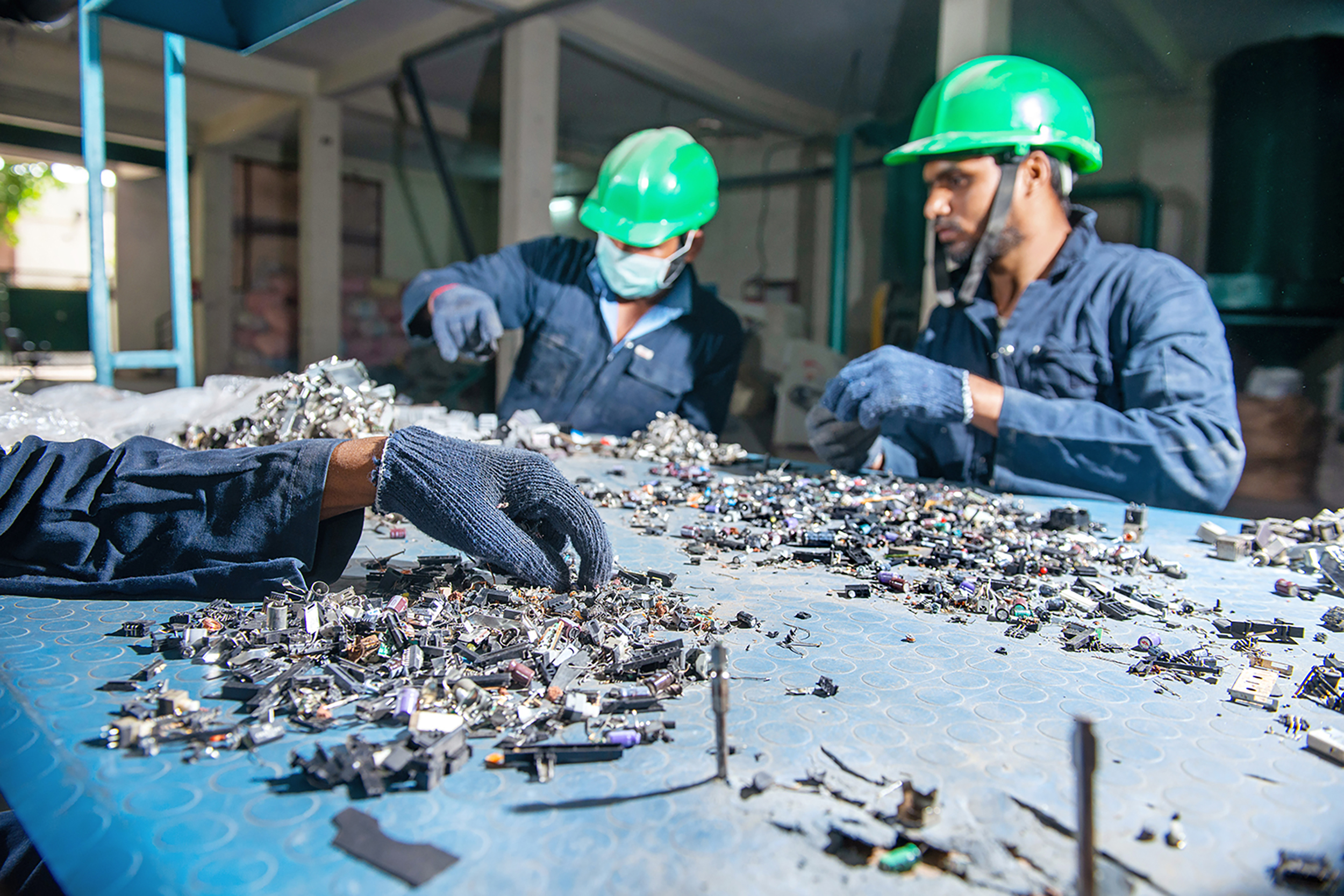 Electronic waste on a shop board with two workers in the background selecting parts for recycling and a hand of a third worker coming from the left side, also selecting parts.