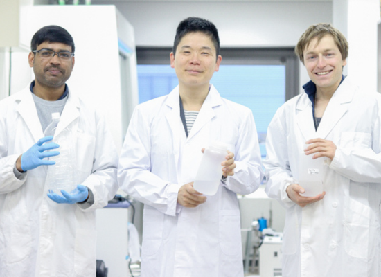three people (indian-, asian- and european-looking) in lab coats holding a plastic container each, smiling into the camera