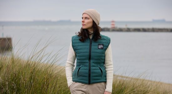 A woman wearing a bodywarmer made from fabrics from the Human Material Loop stands by a lake.