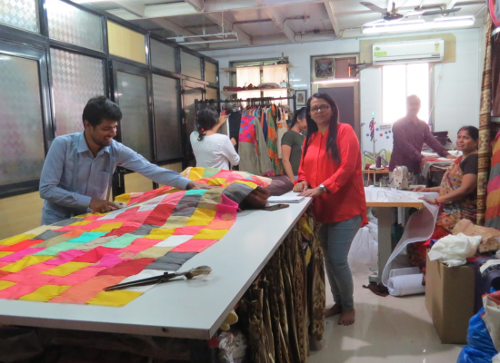 A bright production room. An indian woman in the middle of it with a red shirt smiling at the camera. Several people working on fabrics at different stations.