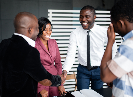 An older, bolder man in a suit is presenting a little machine on a high table to a yound lady in a trade fair booth; both smiling