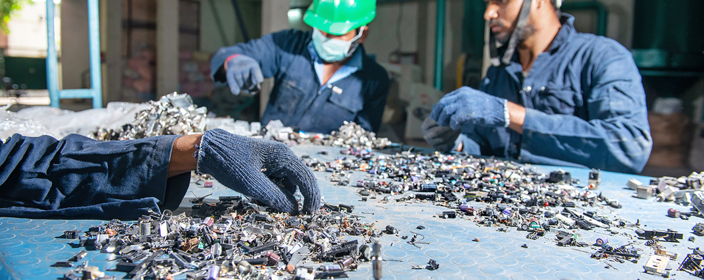 Electronic waste on a shop board with two workers in the background selecting parts for recycling and a hand of a third worker coming from the left side, also selecting parts.